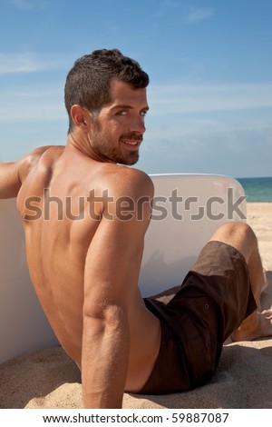 Male surfer sitting on the sand looking back to the camera while holding his board.