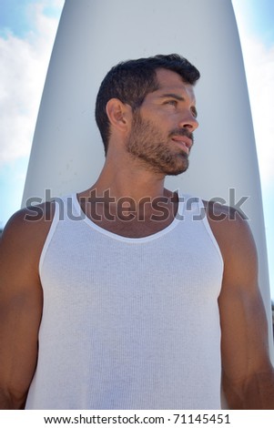 Handsome dark haired surfer wearing a white t-shirt standing in front of his long board with a blue and cloudy sky behind him.