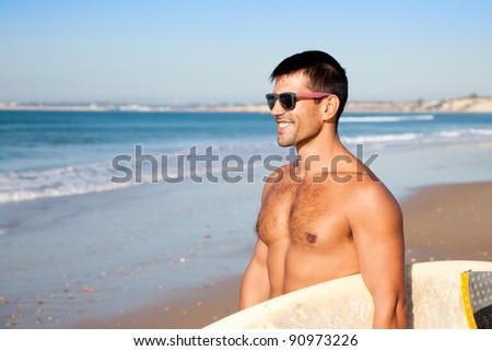Muscular surfer wearing sunglasses smiling and holding his surf board overlooking the ocean.