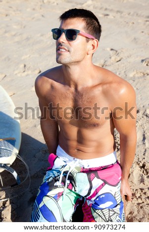 Muscular surfer wearing sunglasses sitting on the sand next to his surf board.