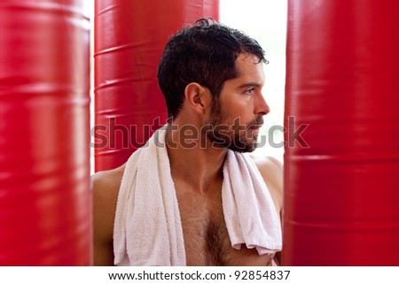 Handsome muscular man with a towel on his shoulders sweating and looking tired in between red punching bags. Fighter.