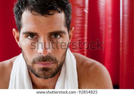 Close up portrait of a handsome muscular man with a towel on his shoulder sweating and looking tired staring at camera in front of red punching bags. Fighter.
