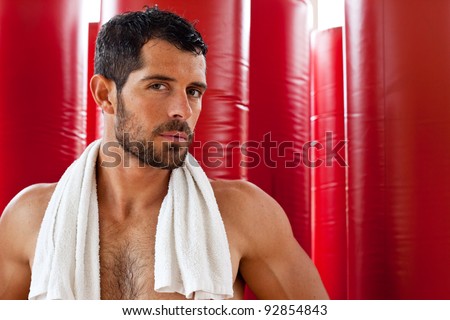 Attractive muscular man with a towel on his shoulders looking at camera in front of red punching bags. Fighter.