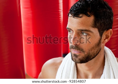 Portrait of a handsome muscular man with a towel on his shoulders sweating and looking tired in front of red punching bags. Fighter.