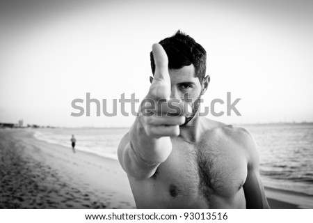 Handsome muscular man on the beach pointing at camera with his hand in the shape of a gun. Black and white portrait.