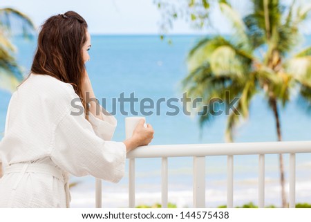 Woman in bath robe looking at beautiful ocean view  from a tropical balcony with morning coffee.