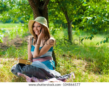 Portrait of a beautiful elegant young woman sitting outdoors barefoot talking on the mobile phone and holding a book in the park or garden on a bright sunny day of summer on the green grass background
