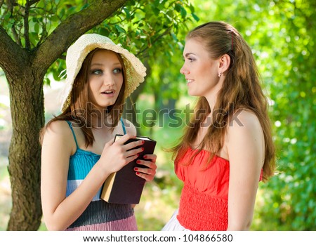 Portrait of a beautiful two young elegant women standing outdoors and one holding a book and talking in the park or garden on a bright sunny day of summer on the green trees background