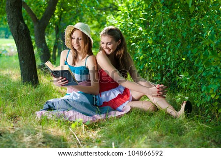 Portrait of a beautiful two young elegant women sitting outdoors reading a book and talking in the park or garden on a bright sunny day of summer on the green grass background