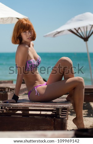 Portrait of a beautiful red haired sexy young woman with a wellness complexion in swimsuit sitting outdoors on bright summer day on beach lounge by blue sea & sky background