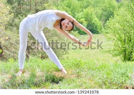Happy smiling young woman doing yoga on green river bank in summer outdoors