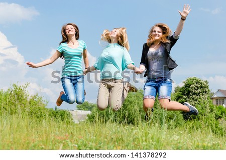 Three happy teen girls friends jumping high on green lawn against blue sky on the bright sunny day & green summer outdoors background