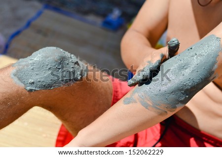Young man om sandy beach applying mineral blue mud on knee and elbow closeup