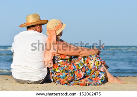 Happy mature couple sitting on sand at seashore having fun outdoors on sandy beach back to back on holiday and looking at sea