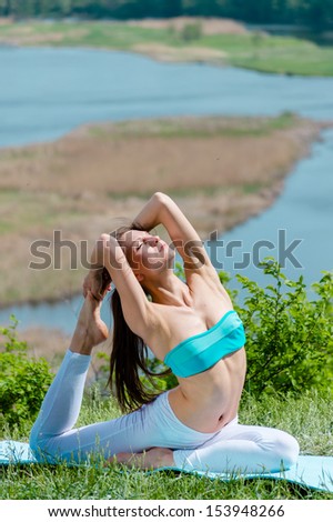 Happy young woman doing yoga exercises in summer outdoors with blue river bank on the green background