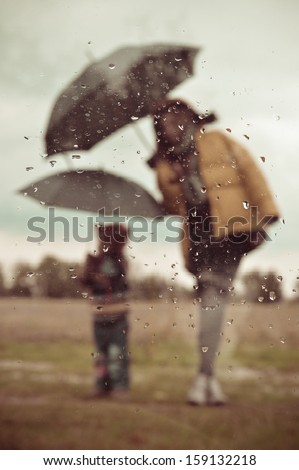 Mother and son under umbrella silhouette through wet window on spring or autumn outdoors background