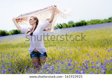 Young smiling woman standing in yellow wheat and bluette field holding a white long piece of cloth in the wind.
