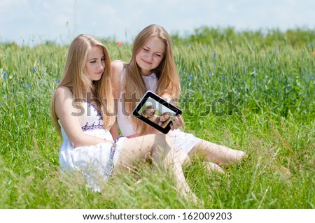 selfshot or selfy portrait: Two happy smiling pretty young women teen girl friends looking on tablet pc & taking picture of themselves while sitting on green lawn on summer outdoors background