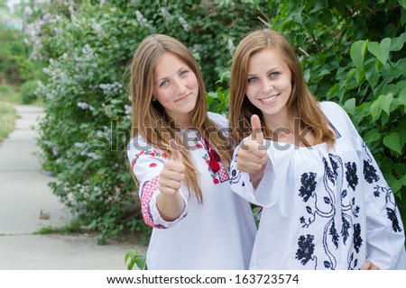 Two young women girl friends giving thumbs up happy smiling & looking at camera on summer outdoors background