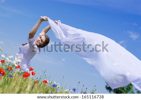 Young smiling woman standing in yellow wheat field holding a white long piece of cloth in the wind on summer blue sky outdoors background