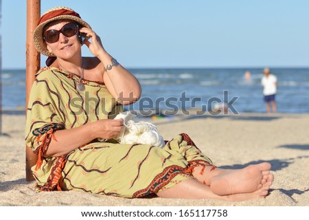 Happy mature woman sitting on beach and talking on mobile phone on summer sea outdoors background