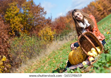 Happy smiling elegant young woman happy smiling & looking at camera with basket of fresh apples sitting on meadow on bright autumn day outdoors background