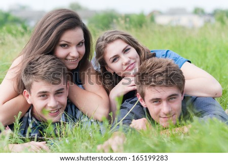 Four happy teenage friends happy smiling & looking at camera lying together on green grass summer outdoors background
