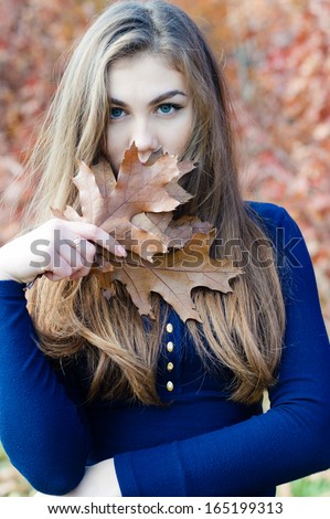 Beautiful elegant woman looking at camera hiding face behind autumn brown leaf on autumn outdoors background
