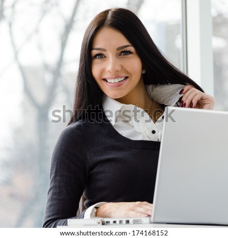 Portrait of a happy smiling & looking at camera young business woman using notebook PC relaxing near office window