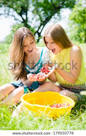 Young beautiful brunette & blond young women girl friends harvested strawberries on summer green outdoors background portrait