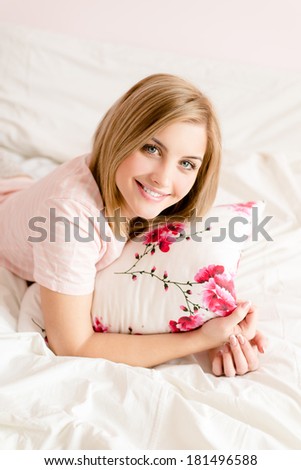 portrait of attractive beautiful happy young blond woman relaxing in bed with floral pillow in hand happy smiling & looking at camera