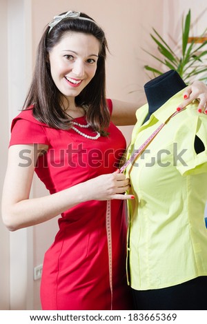 portrait of beautiful funny young pinup woman in red dress with measuring tape & mannequin happy smiling & looking in camera