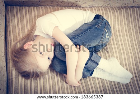 Beautiful child blond girl lying on the sofa hugging knees & looking at camera