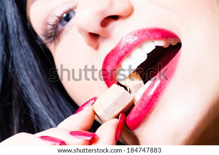 closeup on charming seductive young beautiful woman with red lips and nails, blue eyes biting caramel or chocolate candy portrait