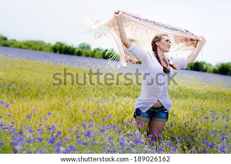 Young smiling woman standing in yellow wheat and bluette field holding a white long piece of cloth in the wind on summer outdoors background portrait