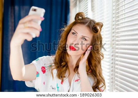 Beautiful young pinup woman taking selfie photo with smart phone at home by window blinds