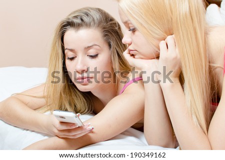 image of 2 adorable blond sisters or beautiful sexy girl friends looking at mobile cell phone having fun relaxing on a white bed