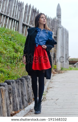picture of beautiful young fashion woman wearing red mini skirt at wooden rustic high fence on autumn day background portrait