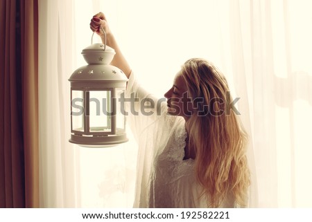 image of beautiful blond young woman holding candle light torch in the early evening & looking at copy space on light window background