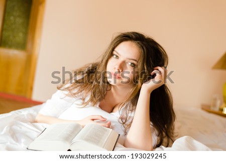 closeup portrait of relaxing charming young sweet brunette woman beautiful girl in white shirt laying on bed reading interesting book & looking at the camera