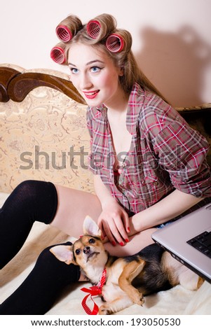 portrait of having fun with little funny puppy pin up girl beautiful blond young woman with curlers on her head with red lips and blue eyes in shirt happy smiling & looking up sitting on bed