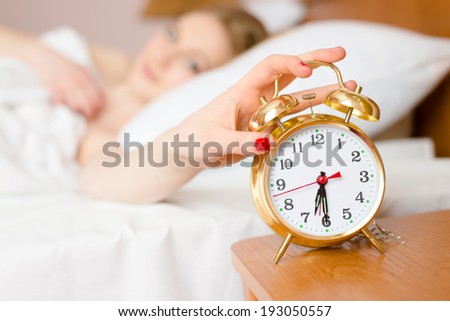 beautiful blond young woman blocking alarm clock off wake up time & looking at clock