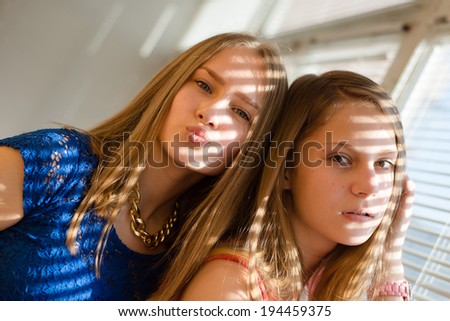 2 blond young women beautiful sisters or girl friends in blue dress having fun posing looking at camera against sun lighted rays through window blinds close up portrait image