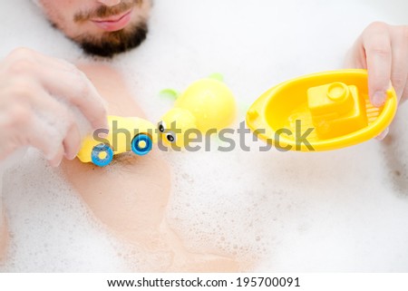 playful male in spa: funny sexy excited man having fun relaxing taking bath tub playing with kid yellow toys in foam holding car & boat in hands close up portrait image