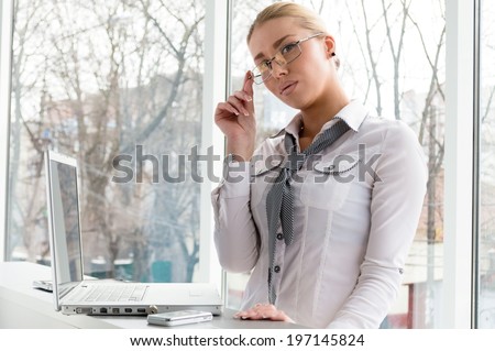 picture of a beautiful blond glamor young business woman using notebook PC computer relaxing having fun seriously looking at camera near office window background portrait