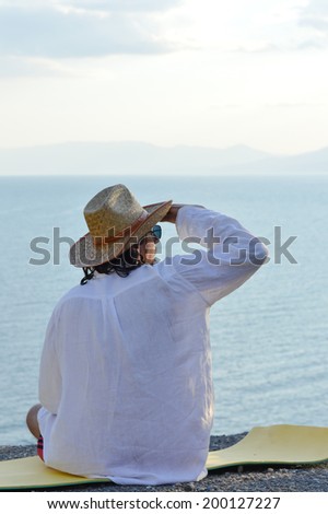portrait of handsome brunette young man in white shirt cool hat & sun glasses having fun relaxing sitting on the sea or ocean beach & looking into the distance on light blue sky copy space background