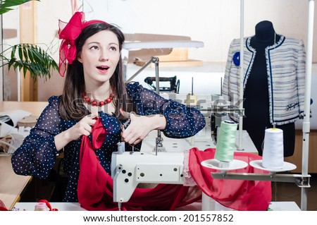 image of dreamy romantic busy working tailoress: beautiful young pinup girl having fun posing with scissors & sewing machine happy smiling looking up at copy space on busy workshop studio background