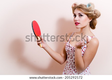 portrait of elegant attractive glamor pinup girl dazedly looking at camera posing in red dress with flower in hair & holding bat ball for table tennis on white or light copy space background closeup