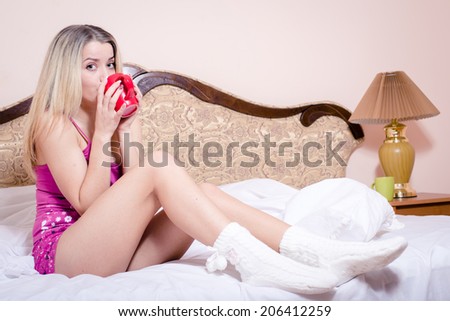 Beautiful happy blond young woman drinking hot tea from red cup sitting relaxing in bed on light copy space background portrait