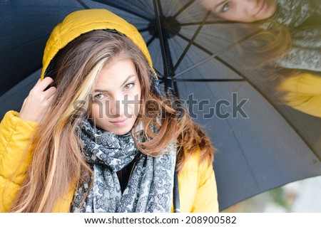 rainy day: closeup on young beautiful brunette woman having fun with umbrella wearing warm yellow coat or puffer on copy space outdoors background, portrait
